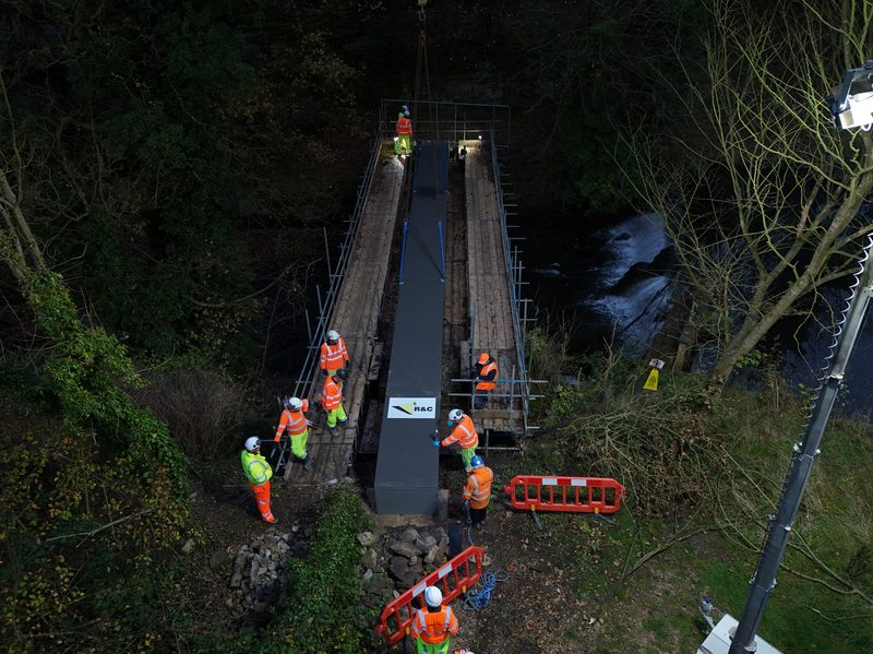 Aerial drone video frame showing hi-vis operatives installing a structural beam over a river at dusk.
