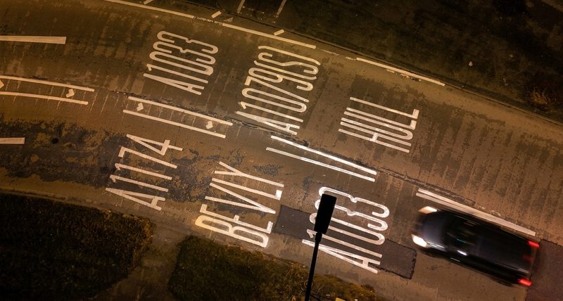 High-angle nocturnal drone view of white road markings and directional signage on a UK dual carriageway.