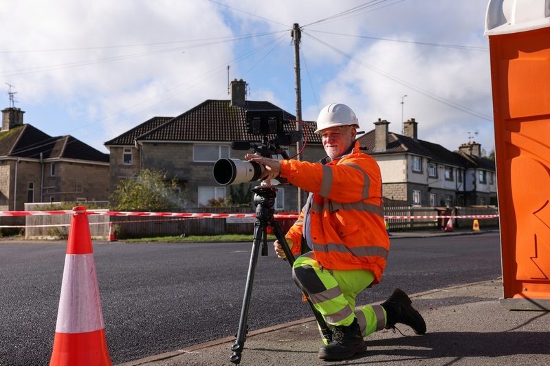 Operative in hi-vis and hard hat filming roadworks with a professional camera on a residential street.