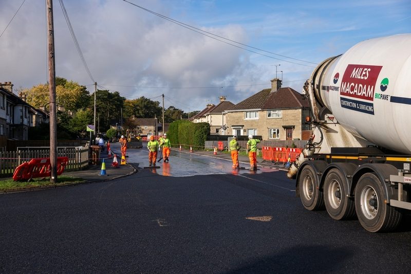Operatives conducting road resurfacing on a residential street with a large material mixer truck and traffic management.
