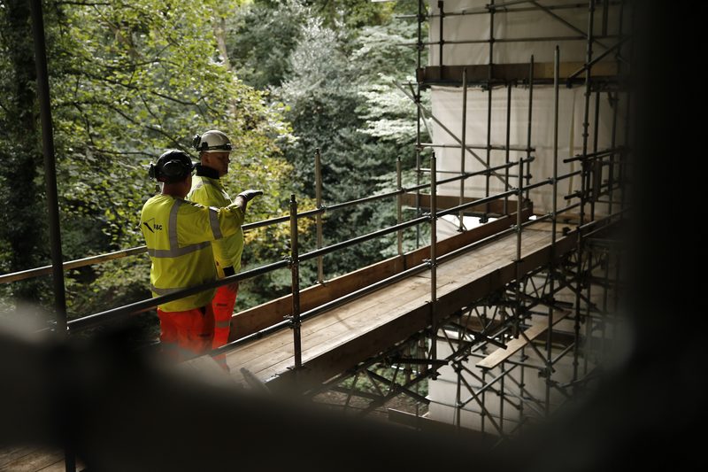 Two site operatives in high-visibility PPE standing on a complex scaffolding structure overlooking a wooded area.