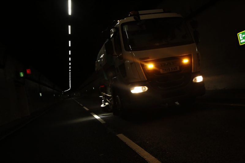 A road sweeper vehicle with amber lights illuminated performing night works inside a dark highway tunnel.