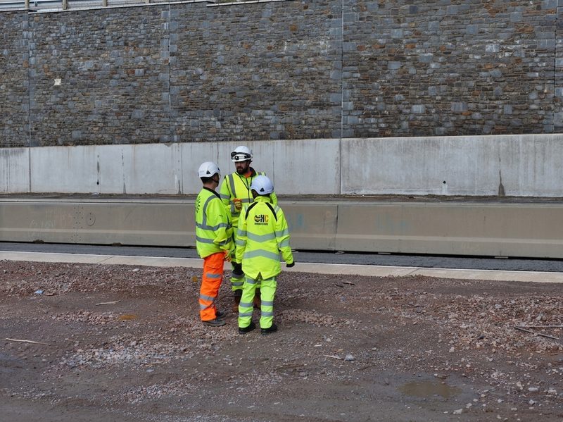 Three highway workers in hi-vis clothing standing in a construction area beside a large stone retaining wall.