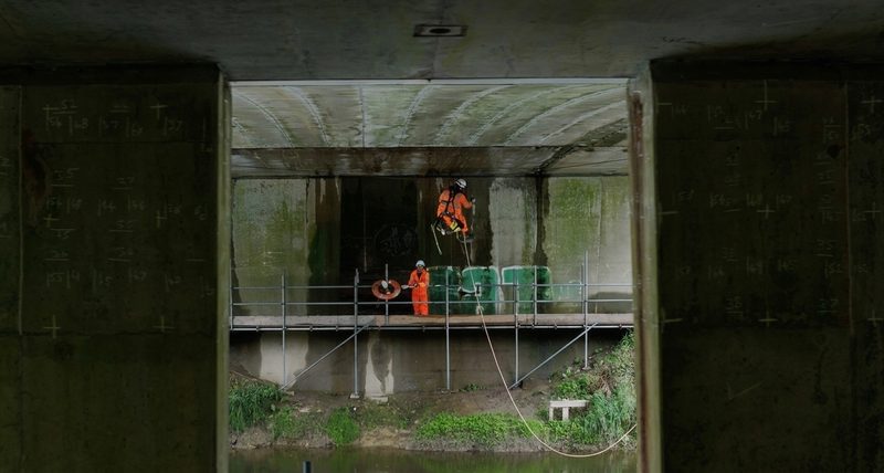 Operatives in hi-vis performing bridge inspections using rope access and scaffolding over a watercourse.