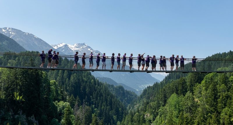 A group of people stand on a high suspension bridge overlooking a forested valley and snow-capped mountains.
