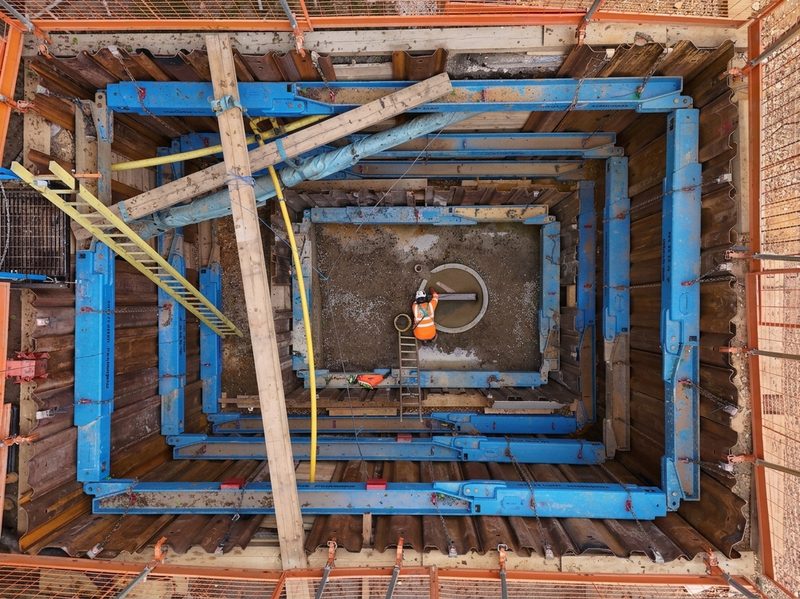 An aerial drone view of a deep shored excavation with steel sheet piling and blue hydraulic bracing.