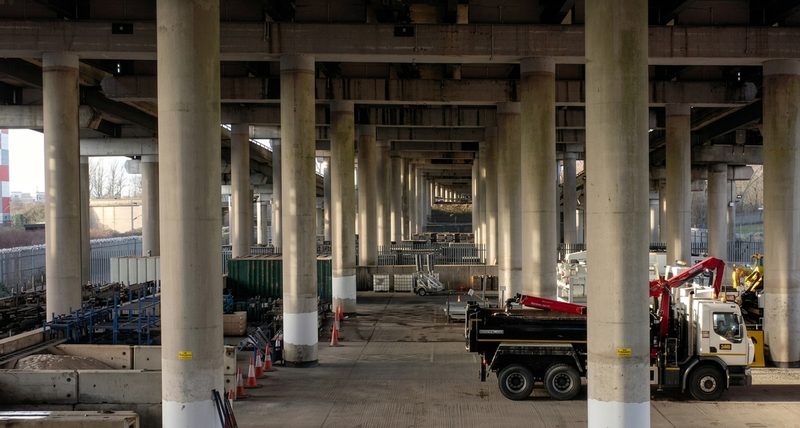 Rows of large concrete columns supporting an elevated highway viaduct with plant machinery and materials stored beneath.