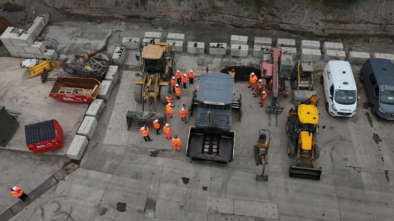 High-angle drone view of hi-vis site operatives gathered for a briefing around highway plant and machinery.