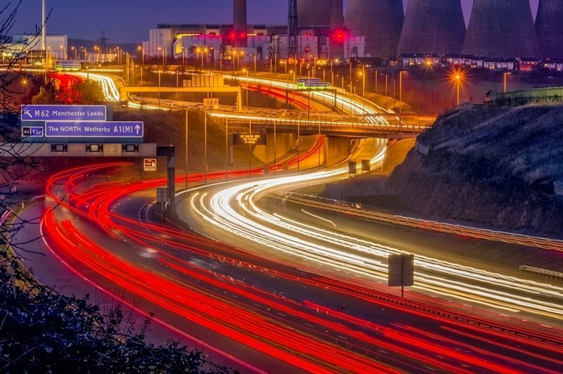 Long-exposure light trails on a busy motorway interchange at night with cooling towers in the background.