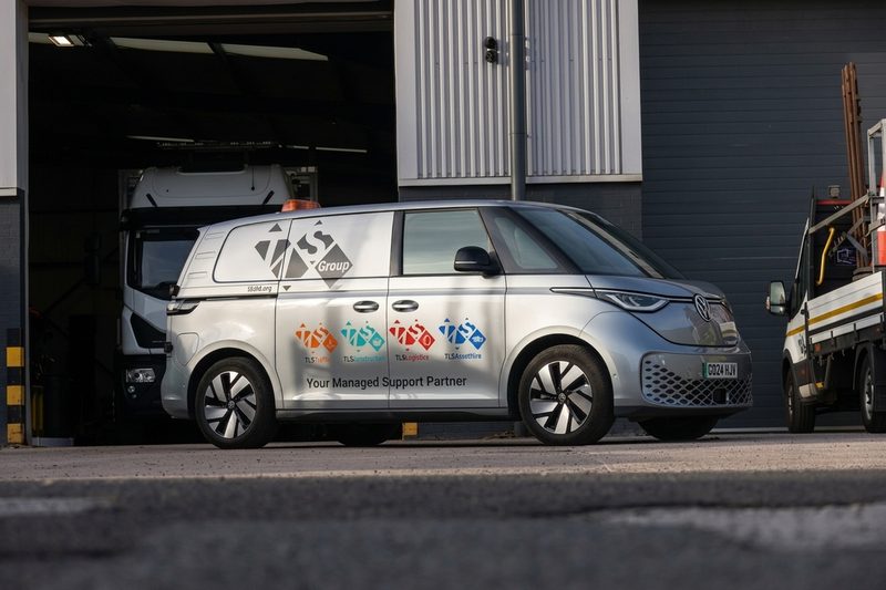 A silver branded electric van parked outside a highways depot with logistics and traffic management logos.