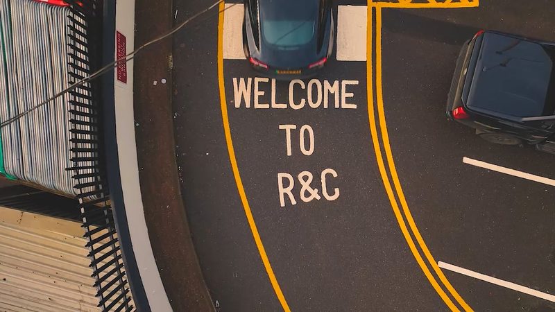 Drone video frame depicting an electric vehicle entering a depot with bespoke road markings and fencing.