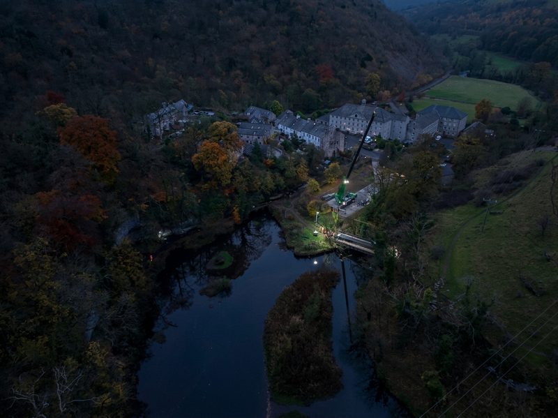 High-angle drone view showing night works with a crane and site lighting near a rural river bridge.