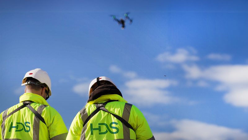 Two highways operatives in high-visibility jackets and hard hats monitoring a drone flying against a blue sky.