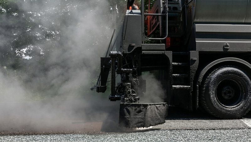 A heavy plant vehicle applying hot bitumen to a road surface with visible steam and vapour.
