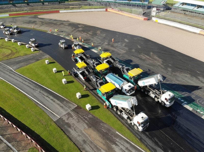 High-angle drone view of multiple pavers and rollers resurfacing a wide section of asphalt track.