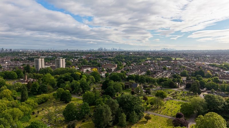 An aerial drone view overlooking green spaces and residential streets with the London skyline in the background.