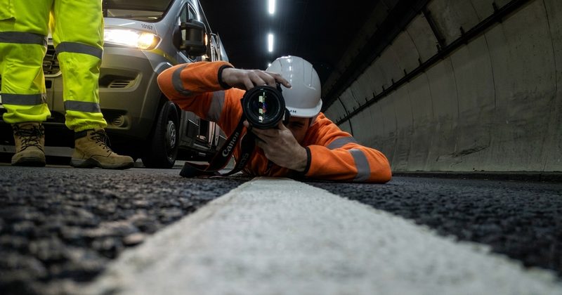 A photographer in high-vis gear capturing a low-angle shot of white road markings inside a lit tunnel.
