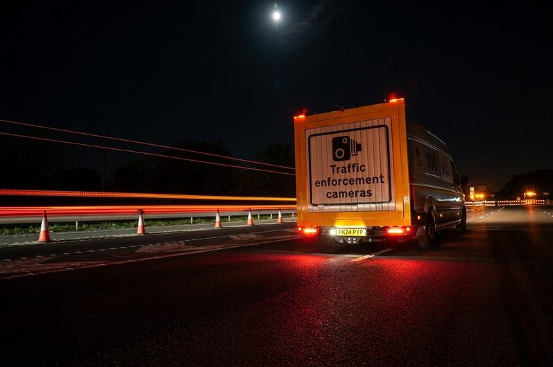 A traffic enforcement camera van parked on a dual carriageway at night with light trails and cones.