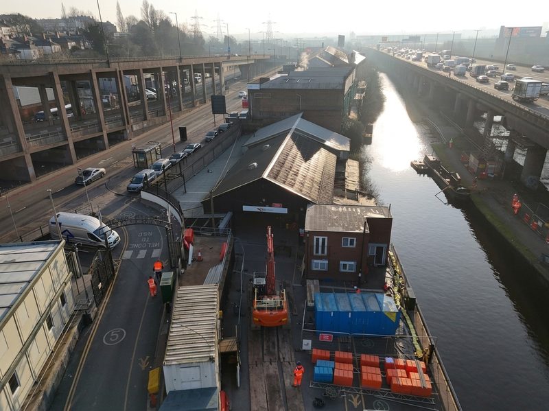 Aerial drone view of a highways depot next to a canal with plant and elevated motorway structures.