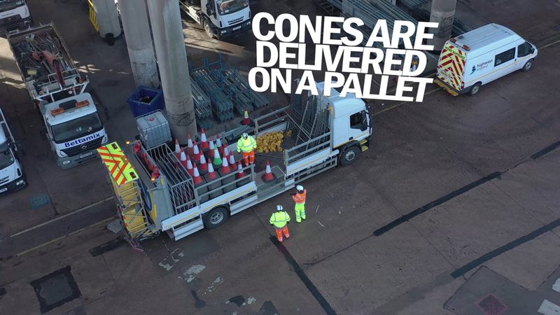 High-angle video frame showing operatives loading traffic cones onto a specialist highway maintenance truck in a depot.