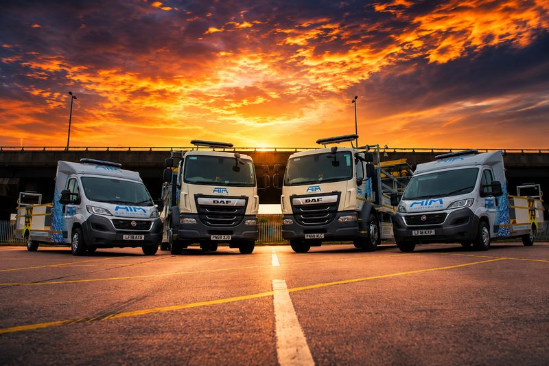 Fleet of highway maintenance vehicles parked in a depot at sunset with a bridge background.