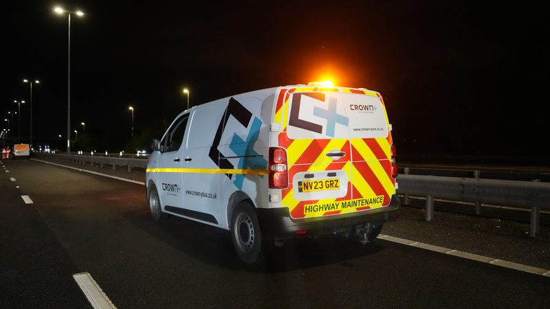 A highway maintenance van with reflective chevrons and amber beacon lit on a dark dual carriageway at night.