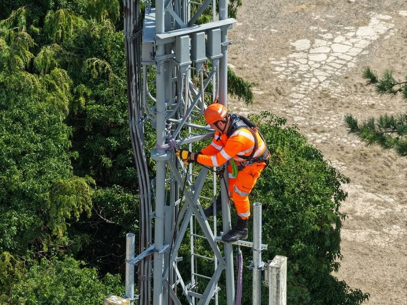 Drone view of an operative in orange hi-vis climbing a steel telecoms mast for maintenance works.