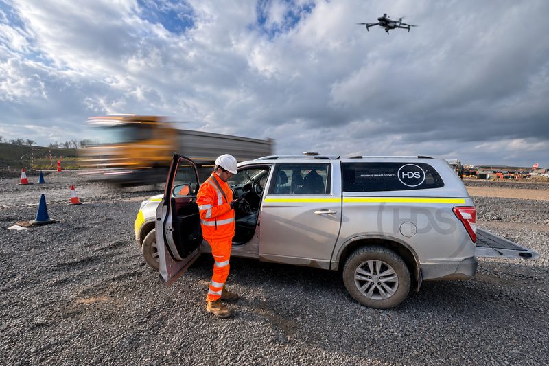 An operative in orange hi-vis stands by a site vehicle whilst a drone flies overhead at roadworks.