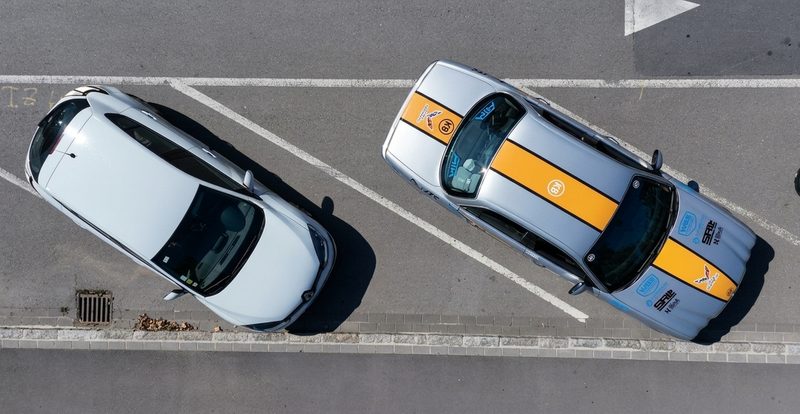 Top-down drone view of two silver cars parked on an asphalt road with white line markings.