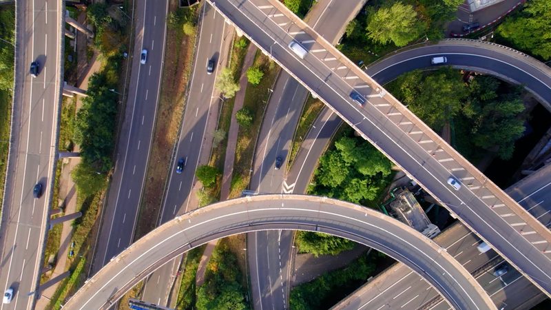 Drone video frame of a complex multi-level highway interchange with moving traffic and greenery.