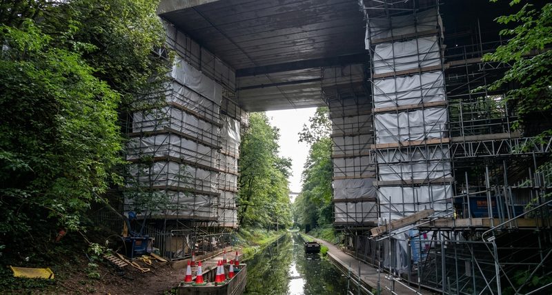 Extensive scaffolding and protective sheeting on a concrete bridge structure spanning a canal in a wooded area.