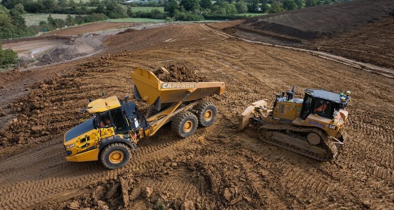 Elevated view of an articulated dump truck and bulldozer performing earthworks on a highway construction site.