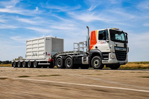 A large heavy goods vehicle with a multi-axle trailer transporting a white industrial container on a highway.