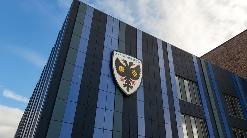 Low angle drone view of the AFC Wimbledon stadium facade featuring blue and black vertical cladding panels.