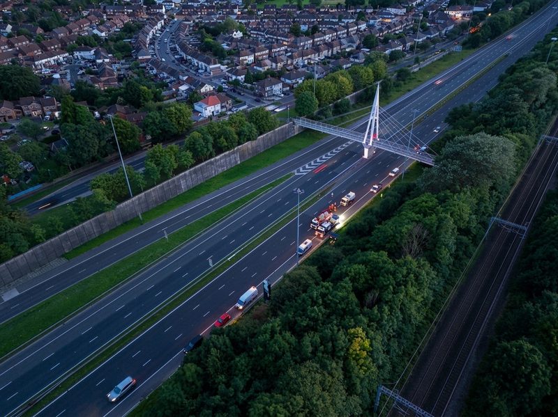 Aerial drone view of motorway maintenance vehicles parked near a lit cable-stayed pedestrian bridge at twilight.