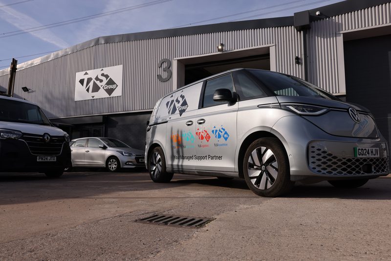 A liveried electric van parked outside a highways depot building with other fleet vehicles in view.