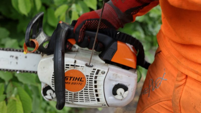 Close-up video frame of a worker in orange hi-vis performing maintenance on a petrol chainsaw.