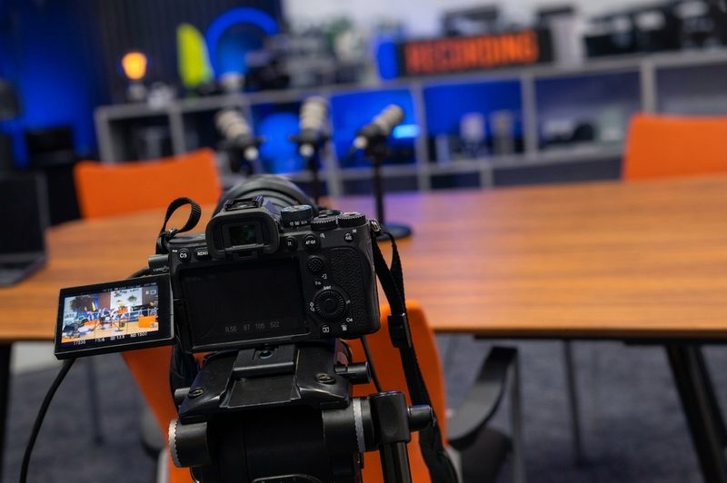 A professional digital camera on a tripod positioned to film a studio recording session with microphones.