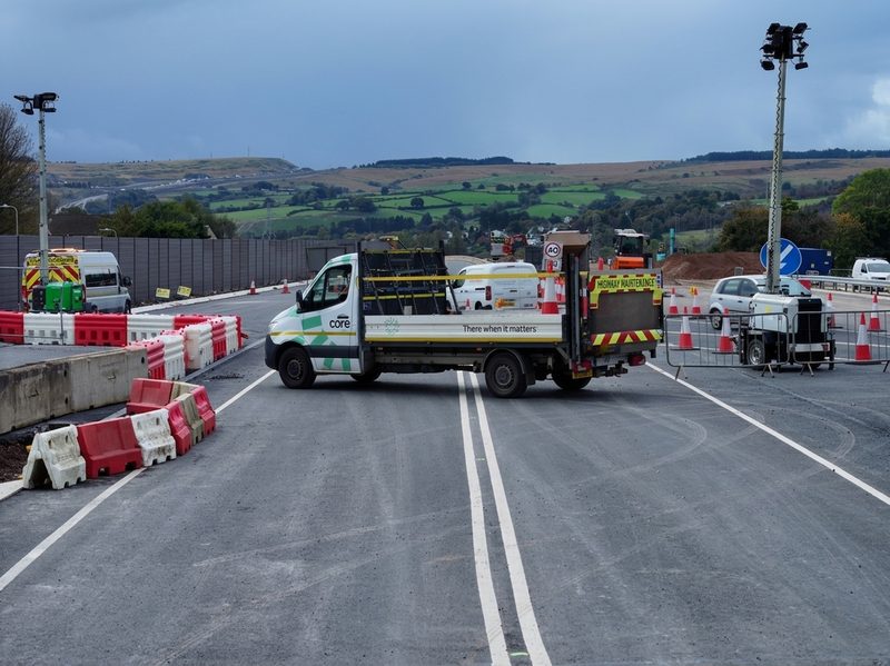 Branded highway maintenance vehicle on a roadworks site with traffic cones, water-filled barriers, and mobile lighting masts.