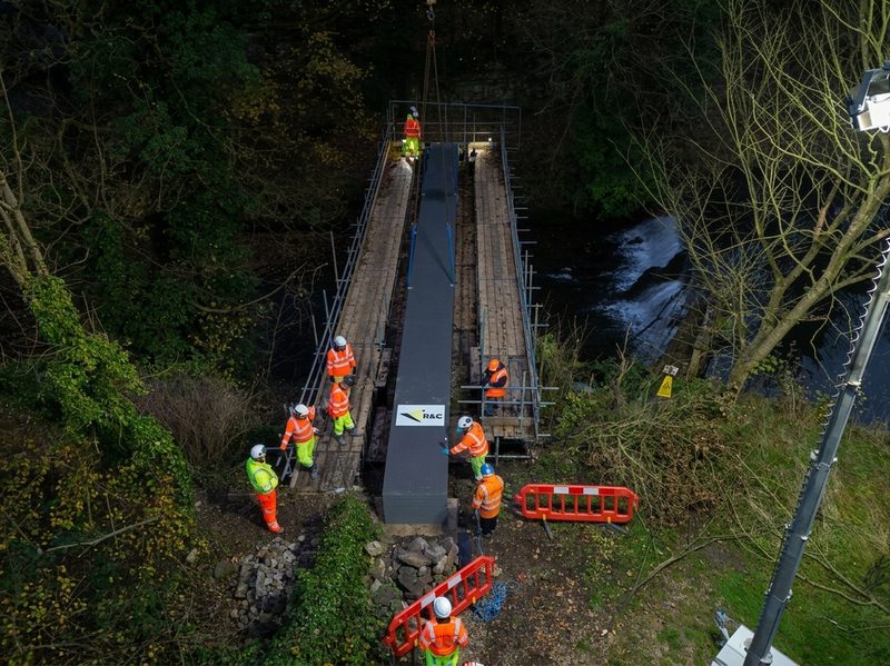 A structural bridge beam being lowered into position by crane with hi-vis operatives on scaffolding platforms.