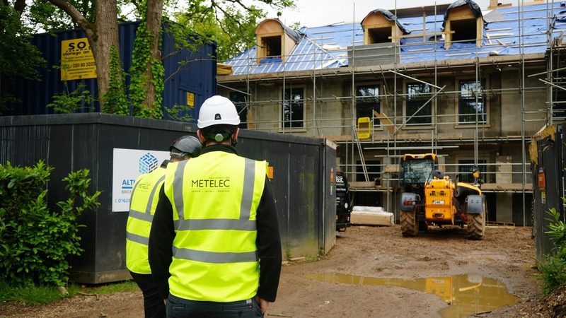 Two operatives in high-visibility vests viewing a building under construction with scaffolding and a yellow telehandler.