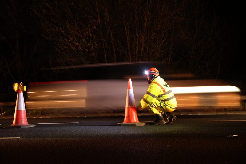 A traffic management operative in hi-vis clothing places cones on a highway during night works.