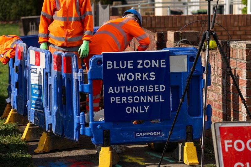 Operatives in orange hi-vis working behind blue pedestrian barriers with signage for authorised personnel.