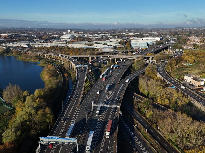 High-angle drone view of a complex multi-level motorway interchange with viaducts, gantries, and heavy goods vehicles.