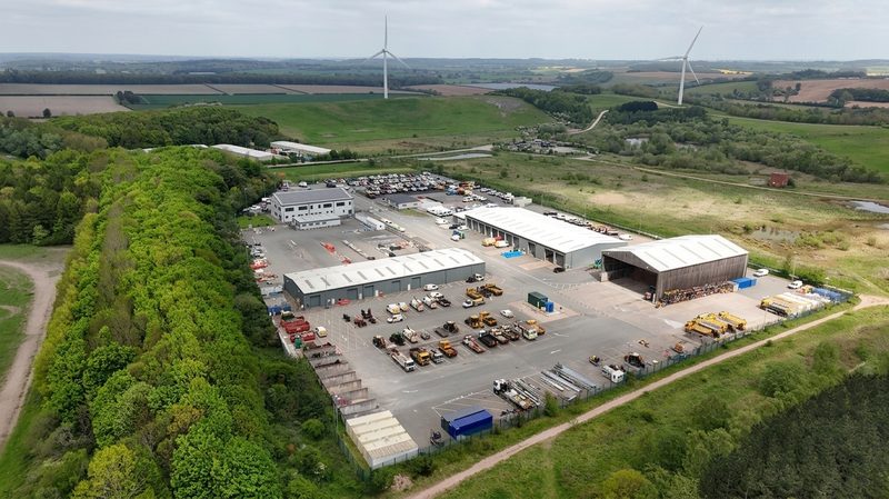 Wide drone view of a highways maintenance depot featuring several large warehouses, parked vehicles and wind turbines.