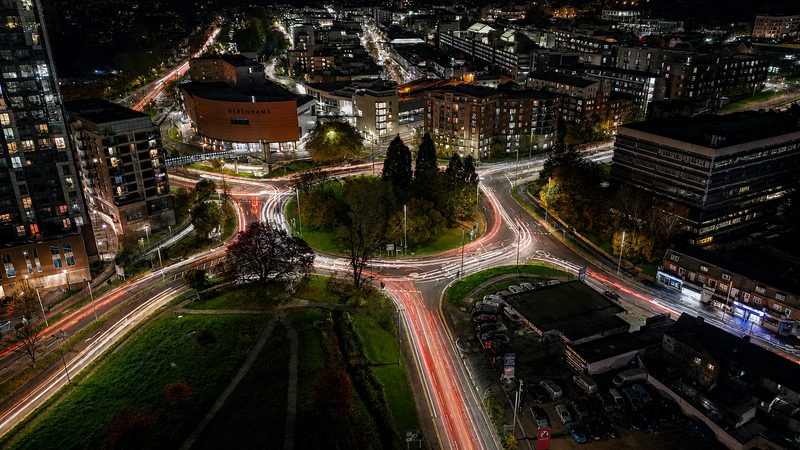 Long exposure drone view of a complex urban roundabout at night featuring light trails from moving traffic.