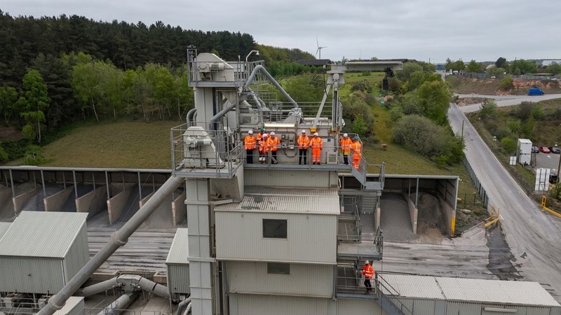 Drone view of operatives in orange hi-vis standing on a high gantry at an industrial asphalt plant.