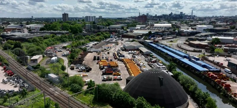 Aerial video frame of a busy highways depot featuring vehicles, railway tracks, and urban skyline.
