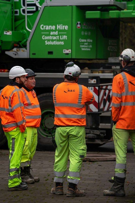 Hi-vis operatives in orange and yellow PPE standing by a green crane during bridge construction works.