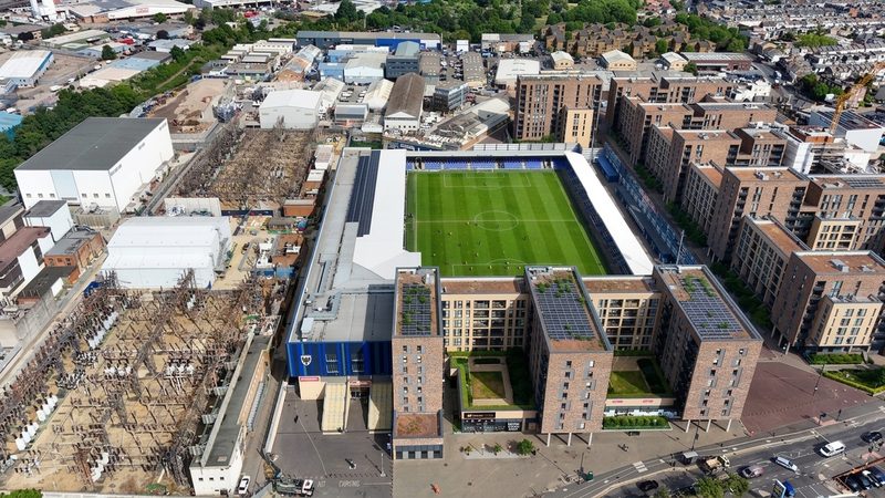 A high-angle drone view of AFC Wimbledon Stadium showing the pitch, surrounding residential blocks, and an electrical substation.